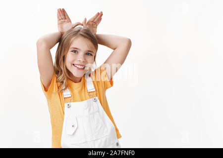 Cute little girl de charme Belle, blonde enfant en t-shirt, bretelles, voir les oreilles de lapin imitant rabit, tenir tête en souriant derrière paumes, jouant joyeusement Banque D'Images