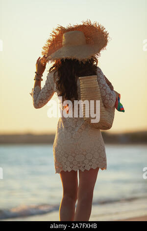 Vus de derrière, l'âge moyen moderne femme en robe blanche et chapeau de paille sur la plage au coucher du soleil la marche. Banque D'Images