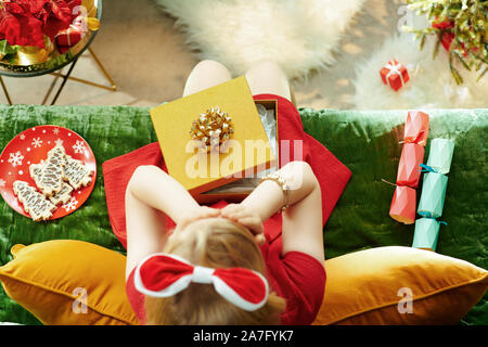 Saison de vacances. Vue supérieure de modern Girl in red dress mécontents de cadeau de Noël tout en étant assis sur le divan dans le salon moderne à Noël. Banque D'Images