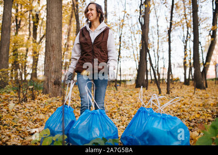 Femme debout à côté d'un tas de sacs poubelles en plastique bleu dans une forêt à l'automne. Banque D'Images