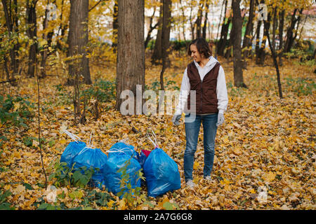 Femme debout à côté d'un tas de sacs poubelles en plastique bleu dans une forêt à l'automne. Banque D'Images