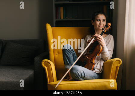 Jolie violoniste femme assise avec un violon à l'aide d'un président à la maison Banque D'Images