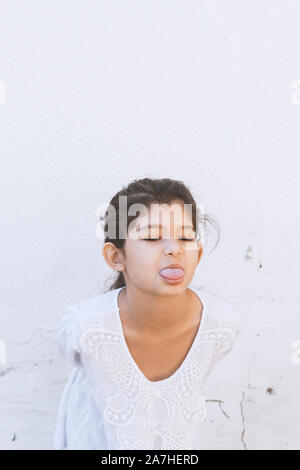 Peu drôle fille en robe blanche montre sa langue. Cute kid girl sticking tongue out. Portrait de jeune femme en colère et malheureux, bouche ouverte et prot Banque D'Images