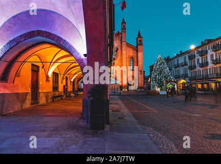 Vue sur les bâtiments illuminés et l'arbre de Noël sur la place pavée en soirée dans la petite ville d'Alba, Piémont, Italie du Nord. Banque D'Images