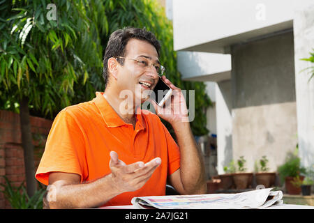 Happy Mid adult male assis-sur-table et Talking On Mobile phone et gesticulant en main-cour près de leur maison Banque D'Images