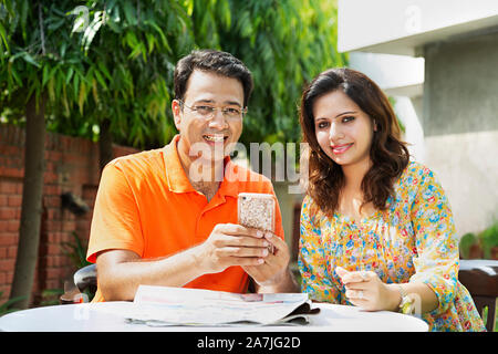 Portrait of mid-adult couple assis-sur-table et en utilisant votre téléphone mobile en-cour de leur maison à Banque D'Images