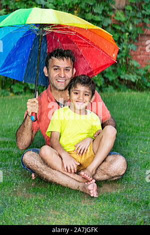 Père de famille heureuse et petit garçon assis-sur-Grass Under umbrella bénéficiant d'Pluie d'été en plein air au parc de la Banque D'Images