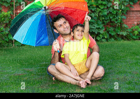 Happy-famille père et enfant garçon assis-sur-Grass avec parapluie du doigt quelque chose dans-la-pluie en jardin extérieur Banque D'Images