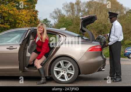 Andover, Hampshire, Royaume-Uni. Octobre 2019. Une passagère blonde portant une jupe courte et un chauffeur habillés, charger des bagages dans le sabot d'une lu Banque D'Images