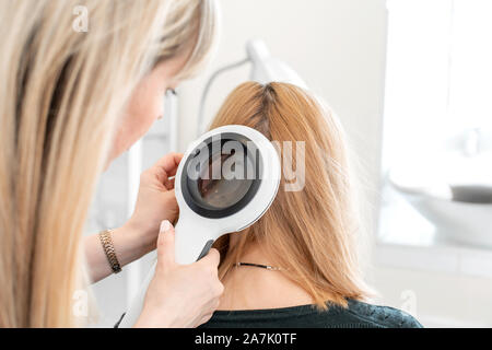 Un trichologist examine l'état des cheveux sur la tête du patient avec un dermatoscope. Dans une salle lumineuse cosmétologie. Banque D'Images