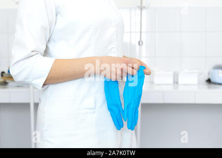 Une infirmière en blouse blanche met sur les gants avant une procédure médicale dans une chambre de traitement lumineux. Banque D'Images