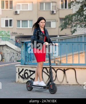 Belgrade, Serbie - 25 octobre 2019 : une adolescente en robe rouge et cuir veste équitation un scooter électrique sur le trottoir de la rue de la ville Banque D'Images