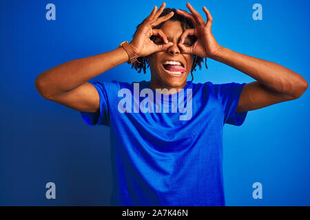 Avec des dreadlocks homme vêtus de t-shirt debout sur fond bleu isolé faisant geste ok comme des jumelles sticking tongue out, les yeux lo Banque D'Images
