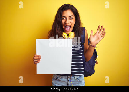 Belle jeune femme étudiante holding banner debout sur fond jaune isolé très heureux et excité, lauréat expression célébrant la victoire sc Banque D'Images