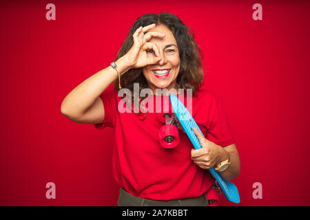 L'âge moyen du patineur senior woman holding skateboard rouge sur fond isolé avec happy smiling face ok faire signer avec la main sur l'œil à par Banque D'Images