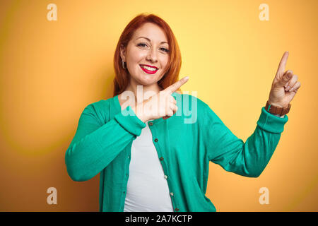 Belle jeune femme rousse portant chandail vert d'hiver isolé sur fond jaune souriant et regardant la caméra pointant à deux mains un Banque D'Images