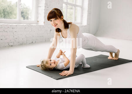 Portrait de belle jeune mère en blanc vêtements sports avec sa charmante petite fille faisant des exercices sur le tapis noir en séance de formation à l'occasion de sport Banque D'Images
