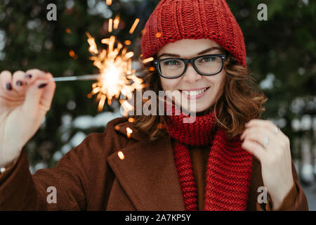 Belle jeune femme en rouge tricoté bonnet et écharpe debout dans le parc avec la lumière du Bengale, Sparkler. Célébration de Noël, concept. L'hiver Banque D'Images