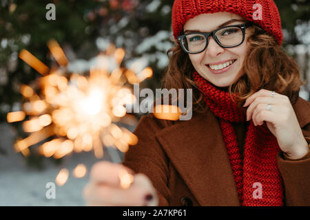 Belle jeune femme en rouge tricoté bonnet et écharpe debout dans le parc avec la lumière du Bengale, Sparkler. Célébration de Noël, concept. L'hiver Banque D'Images