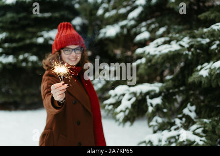 Belle jeune femme en rouge tricoté bonnet et écharpe debout dans le parc avec la lumière du Bengale, Sparkler. Célébration de Noël, concept. L'hiver Banque D'Images