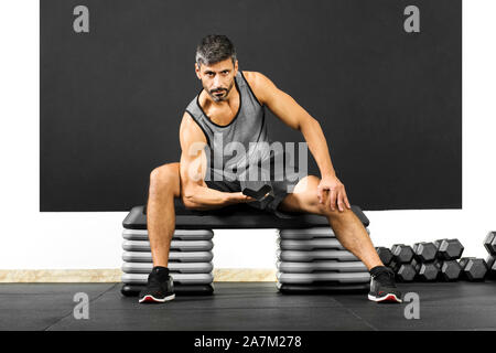 L'homme fit faire avec des boucles un haltère dans une salle de sport assis sur l'étape de l'équipement nécessaire pour renforcer et tonifier ses biceps dans un concept de santé et de remise en forme Banque D'Images