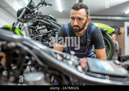Portrait d'un beau motard barbu en salopettes de travail assis près de la moto pendant la réparation dans l'atelier Banque D'Images