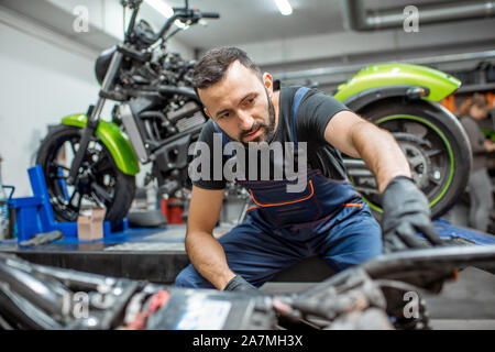 Portrait d'un beau motard barbu en salopettes de travail assis près de la moto pendant la réparation dans l'atelier Banque D'Images