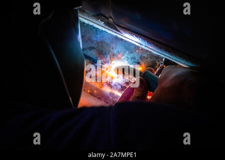 Homme avec un casque et pas de gants de soudure dans le métal d'une voiture et à réparer un trou de rouille Banque D'Images