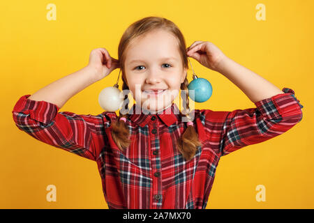 Funny little Girl with christmas ball jouets près des oreilles. Close-up d'un enfant dans une robe rouge sur un fond jaune. Banque D'Images