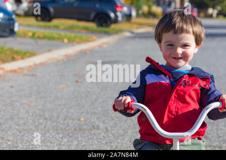 Un jeune enfant, une fillette de 2 ans, monte un tricycle dans une rue de banlieue. Le petit garçon sourit alors qu'il se passe, portant une veste légère pour l'automne ou au printemps, nous Banque D'Images