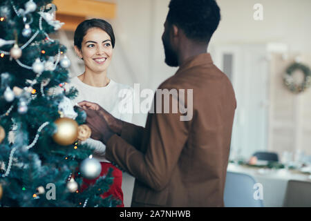 Portrait de race mixte moderne couple decorating Christmas Tree ensemble à la maison, copy space Banque D'Images