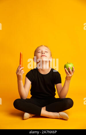 Athletic sept ans girl holding green apple et de carottes à mains isolé sur le jaune Banque D'Images