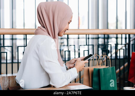 Vue de côté de madame asiatique en hijab smiling et la navigation mobile phone while sitting in cafe près de sacs en papier après le magasinage Banque D'Images