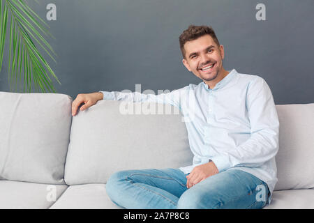 Freestyle. Young man sitting on sofa studio isolé détendue sur joyeux sourire gris Banque D'Images