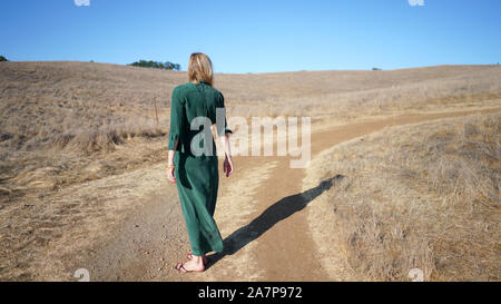 Jeune femme blonde en robe verte de longue date à l'opposé de l'appareil photo d'une grande ombre sur chemin de terre dans un sol sec, pays Banque D'Images