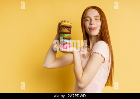 Shot of woman with red hair, fermé les yeux, léchant ses lèvres avec la langue, tenant dans les mains de délicieux donuts colorés plus isolés yello Banque D'Images