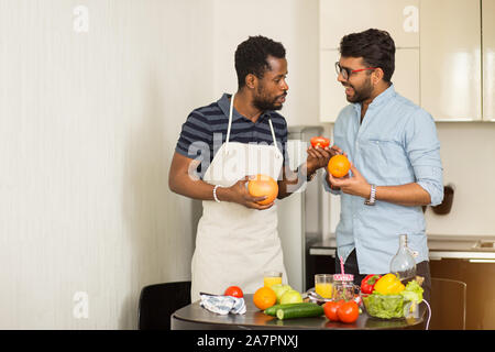 Deux beaux mâles joyeux amis debout dans la cuisine de fruits dans les mains, parler, faire du jus d'orange et de pamplemousse, la préparation des aliments sains t Banque D'Images