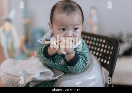 Bébé asiatique assis sur une chaise à l'theeting avec toy banane rayé et visage blessé de sa main à la maison Banque D'Images