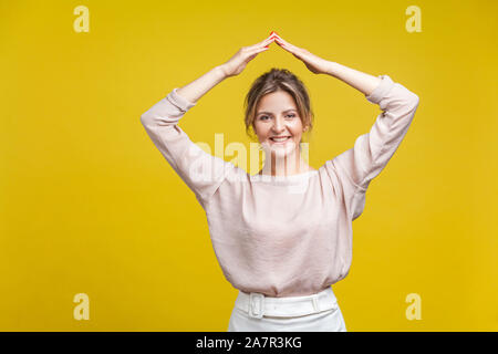 Je suis en sécurité. Magnifique Portrait de femme sympathique avec les cheveux clairs en Chemisier décontracté debout avec les bras sur la tête, montrant le geste de toit, symbole accueil. je Banque D'Images