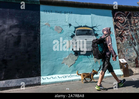 Mur de Berlin graffiti East Side Gallery Allemagne jeune homme marchant Le chien qui passe autour de la voiture Trabant Friedrichshain City Street Berlin Les gens de la ville Banque D'Images