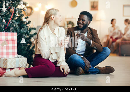 Portrait de race mixte couple clinking glasses champagne assis par arbre de Noël à la maison, copy space Banque D'Images