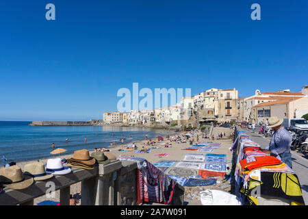 Un homme de vendre ses marchandises sur le front de mer de Cefalú, Sicile, Italie, comme beaucoup de gens sur la plage profiter du beau temps. Banque D'Images