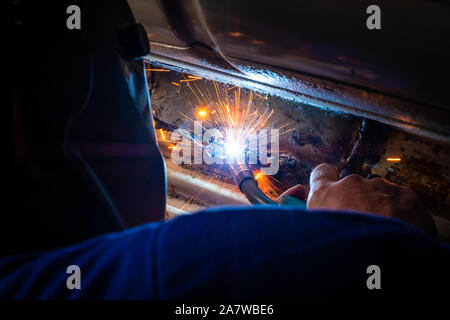 Homme avec un casque et pas de gants de soudure dans le métal d'une voiture et à réparer un trou de rouille Banque D'Images