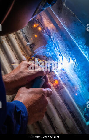 Homme avec un casque et pas de gants de soudure dans le métal d'une voiture et à réparer un trou de rouille Banque D'Images