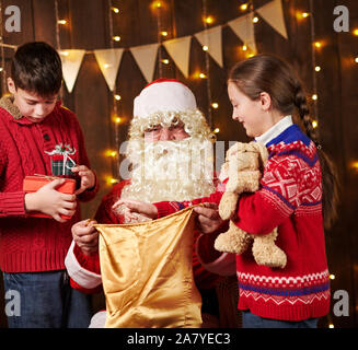 Le Père Noël ouvre un sac et donne des cadeaux aux enfants, assis près de l'arbre de Noël décoré avec des lumières - Joyeux Noël et bonnes fêtes ! Banque D'Images
