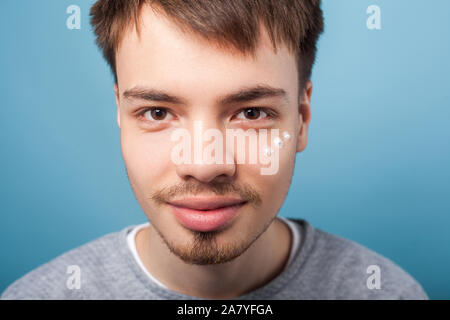 Closeup portrait of positive brunette homme avec petite barbe smiling at camera, avoir des taches de crème hydratant sous les yeux, l'application de traitement du visage. Banque D'Images