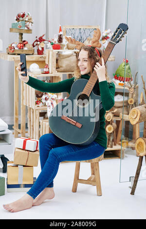 Young cheerful woman wearing reindeer antlers quand posant avec guitare la veille de Noël Banque D'Images