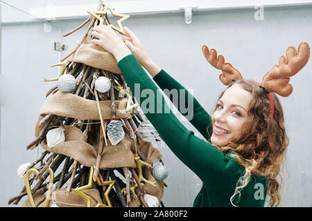 Cheerful young woman in reindeer antlers decorating Christmas Tree at home Banque D'Images