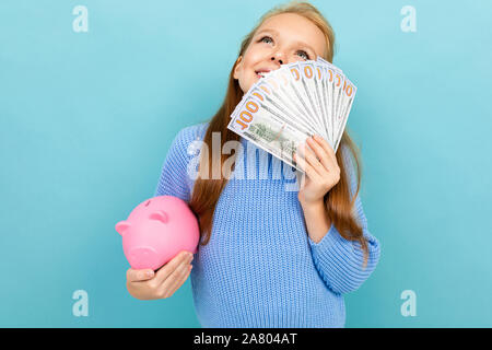 Rêver cheerful girl lycéenne contre un mur bleu avec une tirelire et un ventilateur d'argent Banque D'Images