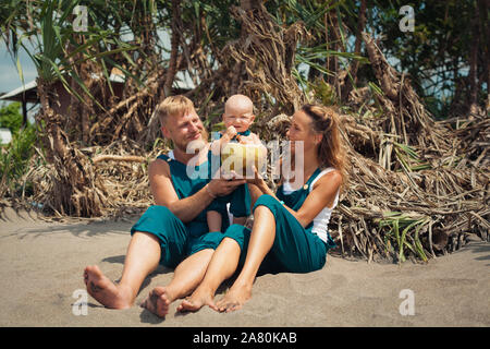 Drôle de famille heureux hipster pique-nique sur plage - mère, père nourrir bébé garçon. Petit-fils de manger des fruits avec plaisir, boire de noix de coco fraîche. Mode de vie sain Banque D'Images